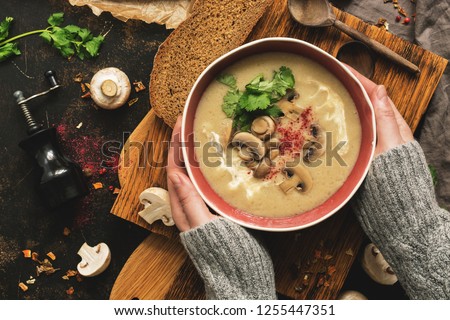 Bowl of mushroom cream soup in woman hands in a woolen sweater. Rustic dark background, top view, flat lay. Winter warming soup on vintage cutting boards