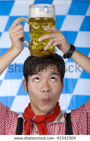 Asian Boy, dressed with Bavarian Lederhose holds an full Oktoberfest beer stein on his head and looks up. In background is the Bavarian flag visible.