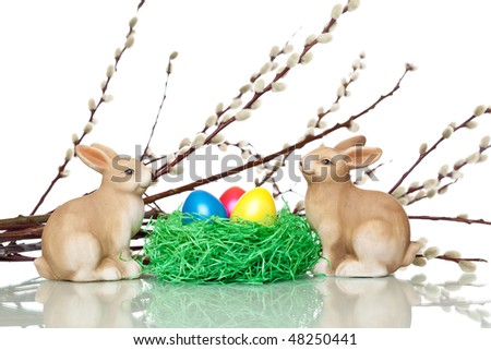 Two cute Easter bunnies sitting beside an Easter nest. In the nest is a yellow, a red and blue Easter egg. In background is a catkin (pussy willow) visible.