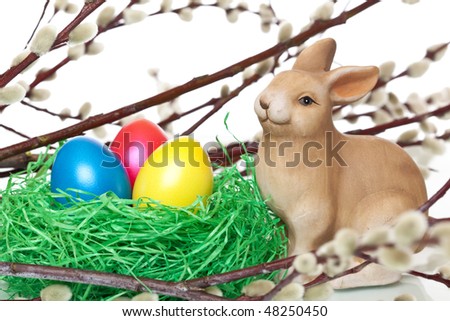 One cute Easter bunny is sitting beside an Easter nest. in the nest is a yellow, a red and blue Easter egg. In background is a catkin (pussy willow) visible.
