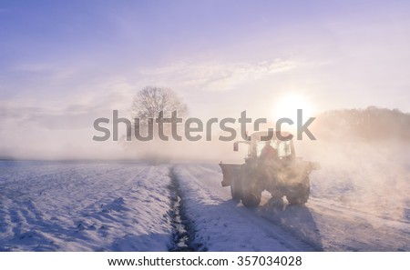 Misty Winter landscape with a countryroad, warmed up by sunrise and  wrapped in a coat of fog. From the mist it can be seen a silhouette of a farmer driving its tractor on a frosted field.