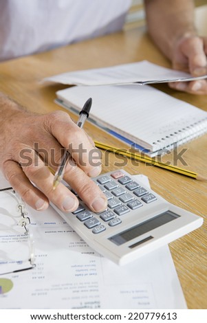 Close-up detail of older man\'s hand holding a pen and using a calculator with paperwork in background