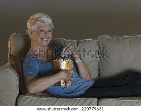 Evening scene of mature woman lounging on sofa watching TV and eating popcorn