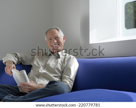 Relaxed portrait of a senior man sitting with a book looking to camera in a contemporary interior