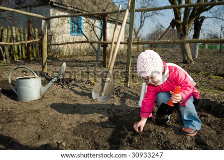 An anxious little girl squatting down on the ground cultivating soil with a toy spade