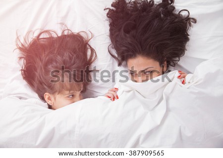 Portrait of brunette long hair mother and daughter under the duvet together in soft morning light on white linen bed. Concept of happy family living, relaxation, comfort, fun. Top view.