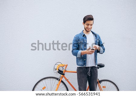 Young student standing near the orange bicycle looking at phone and listening to music. Attractive man with beard is trying to find his the best track during the stop. He is glad of doing this. Wall
