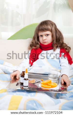 Sympathetic little sick girl in red scarf under blanket in bed is taking a salver with pills, sprays and orange