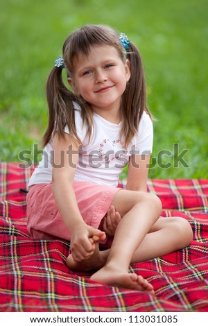 Little smiling cute blond girl preschooler with ponytails sitting on the red plaid on green grass in summer