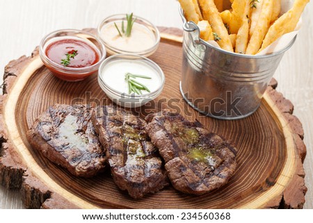 Portion of BBQ beef filet mignon steak  served  on wooden board with  ketchup, mustard and cream sauces, fried potatoes in aluminium bucket