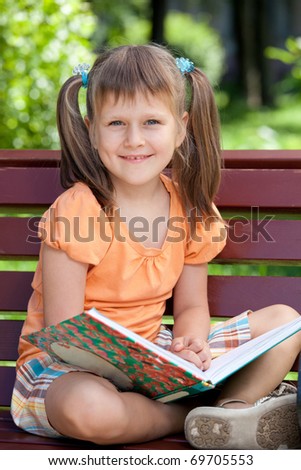 Portrait of little cute smiling girl preschooler with open book who is sitting cross-legged on the wooden bench in summer park