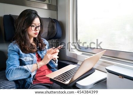 Cute Asian woman using smartphone and laptop on train, copy space on window, business travel or technology concept