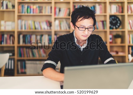 Young pensive Asian man working on laptop at home office or library with serious face, bookshelf with clock blur background with copy space, business or technology concept