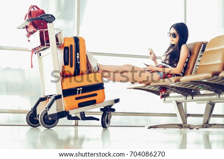 Young Asian traveler woman, university student sit using smartphone at airport, luggage and bag on trolley cart. Online check in mobile app, study abroad, or international tourism lifestyle concept