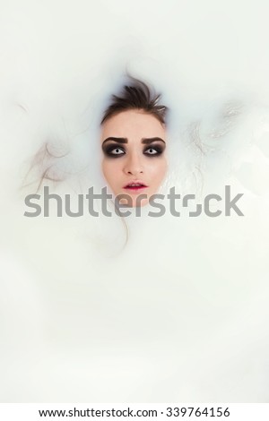 Scary looking smoky eyes brunette girl face with hair under milk. Halloweet portrait in bath with white milk background.