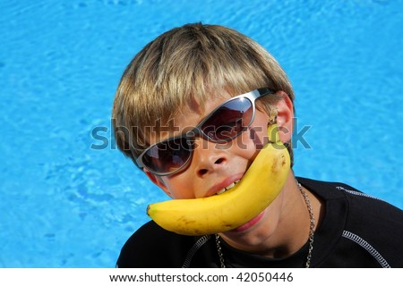 a 10-years old American - German boy with sunglasses joking with a banana in his mouth sitting at a swimming pool in the summer sun
