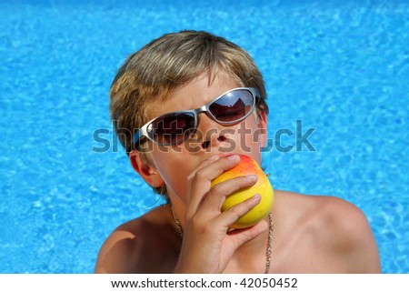 a 10-years old American - German boy with sunglasses sitting at a swimming pool and enjoying eating a delicious apple with closed eyes in the summer sun