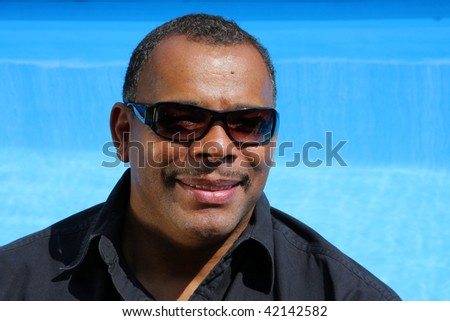 a smiling, successful and happy African-American man with sunglasses photographed in the summer sun in his garden sitting at his swimming pool