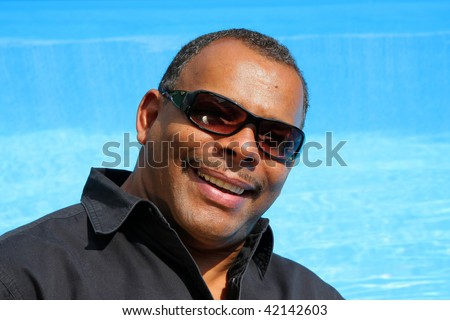 a smiling, successful and happy African-American man with sunglasses photographed in the summer sun in his garden sitting at his swimming pool