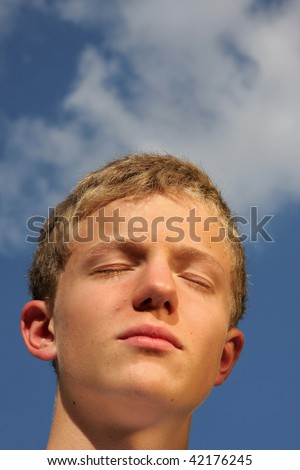 a handsome, smiling and happy 14-years old sun tanning teenager with closed eyes photographed in the summer sun with blue sky and clouds in the background