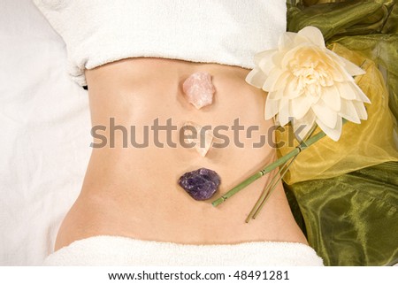 a wellness composition with a closeup of an abdomen of a natural mature woman with stones, flower and a silk scarf