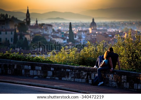 A couple in love - girl and boy sitting on a small wall by the road watching a scenic sunset over a romantic Italian city on the hills in the blurred background; in Florence, Italy