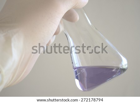 Closeup of an Erlenmeyer Flask filled with a blue liquid or clear water, held by a gloved hand in a lab, a hospital or some other laboratory environment
