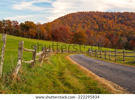 An old country road lined with a split rail fence turns as the autumn leaves glow with color against the North Carolina sky.