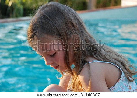 Young girl sitting in the border of the swimming pool, smiling. Her blond hairs covers her shoulders, she is in very peaceful mood.In the background there is  a blue water of the swimming pool