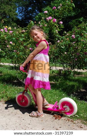 Little girl on the child\'s scooter in the in the park. Her dress and scooter have rose and white colours, and in the background there are roses of similar colours