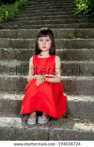 Charming little brunette girl in a red dress sitting on stairs