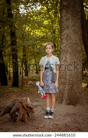 Charming sad little girl standing near  tree in forest with  doll in hands