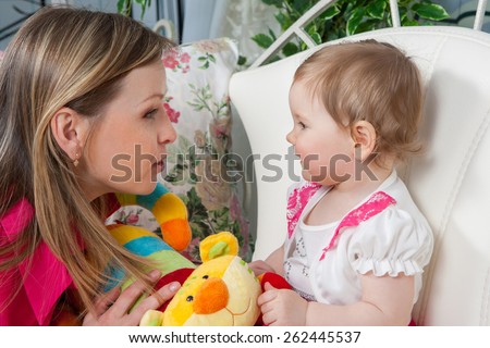 Happy mother with  baby daughter in a beautiful dress at home near the fireplace. Mom tells daughter story. Baby listens with delight.