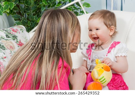 Happy mother with  baby daughter in a beautiful dress at home. Mom tells daughter story. Baby listens with delight.