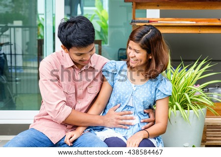 young adult asian  husband touching his pregnant wife belly on sofa, moving to new home, new life of asian couple parent with pregnant baby at new home start, smiling happy asian family