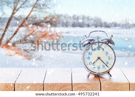Vintage alarm clock on wooden table or bench in snowy weather on winter background. Return to winter time. Fall back time. Daylight savings end. Switched to winter time changing clock from wintertime.