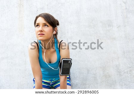 young woman resting after run by the wall in city with big smile. in blue sportswear. listing music.