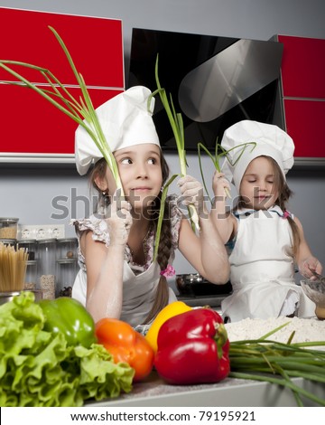two little girls in clothes cooks in the kitchen at the table with raw food, with bunches of green onions in the hands of