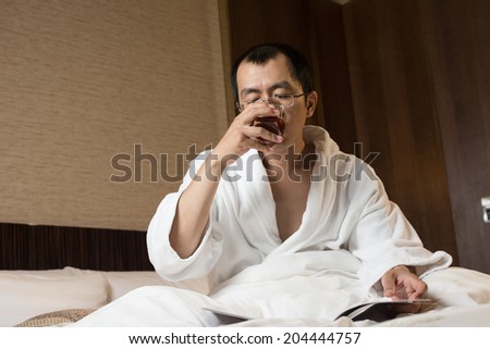 Mature Asian man in bathrobe in hotel, reading magazine.