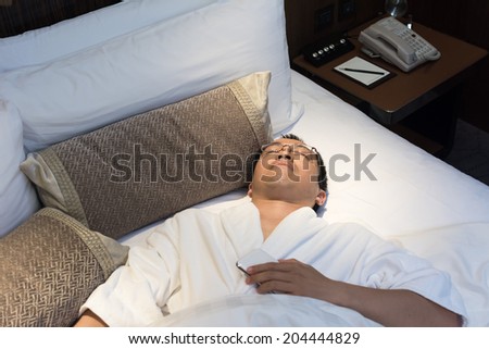 Mature Asian man in bathrobe in hotel, sleeping on bed.