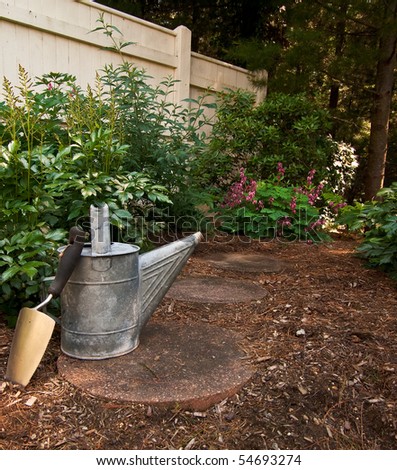 A trowel sits against an old watering can on a stone path that leads to a garden in the woods
