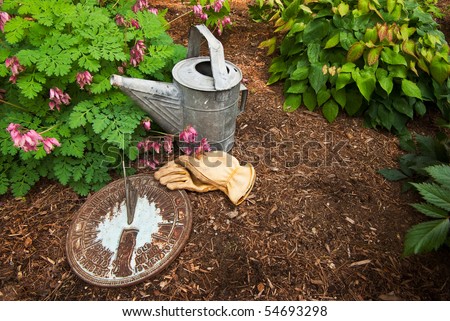 An old sundial sits in a garden next to a worn watering can and leather work gloves