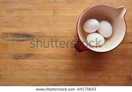 A view looking down on a red ceramic bowl filled with eggs on a butcher block counter