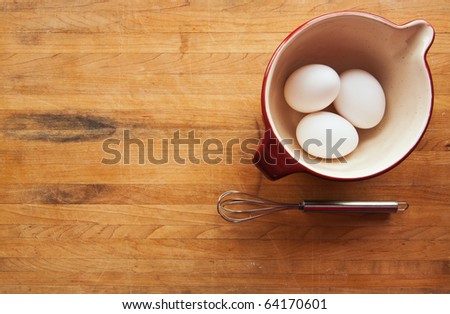 A view looking down on a red ceramic bowl filled with eggs on a butcher block counter with a whisk sitting to the side