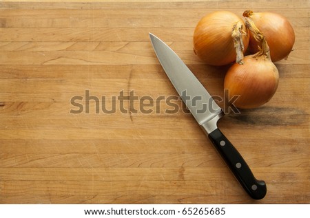 A view looking down on a group of onions and a large knife on a worn butcher block cutting board