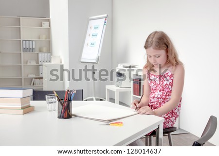 Pretty young girl in a floral summer frock sitting on a stool at a table drawing on a large sketchbook alone in a classroom or home office