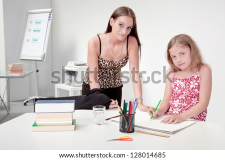 Mother or teacher supervising a pretty little girl as she sits at a table with a large sketch book drawing, interior portrait in an office or classroom