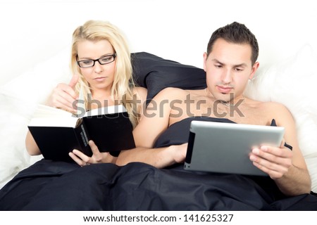 Married couple reading propped up against the pillows before sleeping with the wife reading a heavy novel while her husband enjoys an e-book on his tablet