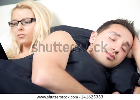 Closeup of an attractive young man sleeping in bed with his wife busy reading behind him propped up against the pilows