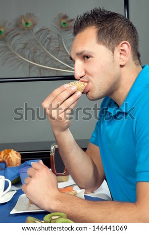 Young man eating breakfast biting into a fresh roll while seated at a table set with fresh fruit and coffee